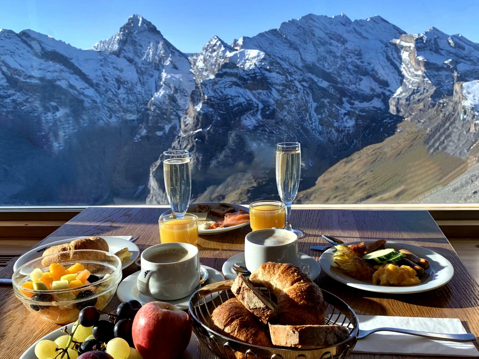 Plates of brunch and glasses of prosecco on a table at Schilthorn Piz Gloria restaurant, overlooking a mountain.