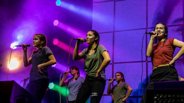 Two rows of young women stand with their hand on their hips, holding a microphone as they sing on a purple lit stage