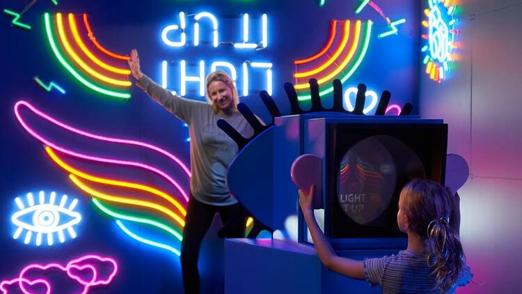 A woman stands in front of a neon wall of rainbow colours, her child takes a photo of her