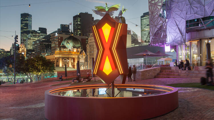 A circular pond, with a glowing sculpture in an X shape above it, is lit up at night, the backdrop is Fed Square and Melbourne city