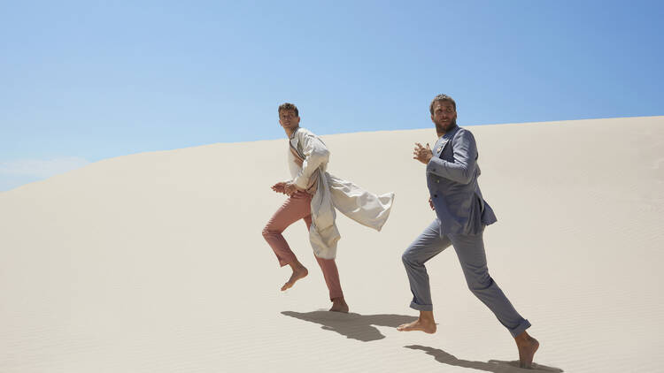 Two men in suits run barefoot on a sand dune.