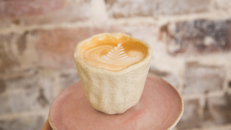 A coffee in a clay cup sits on a pink pottery plate against an exposed brick wall