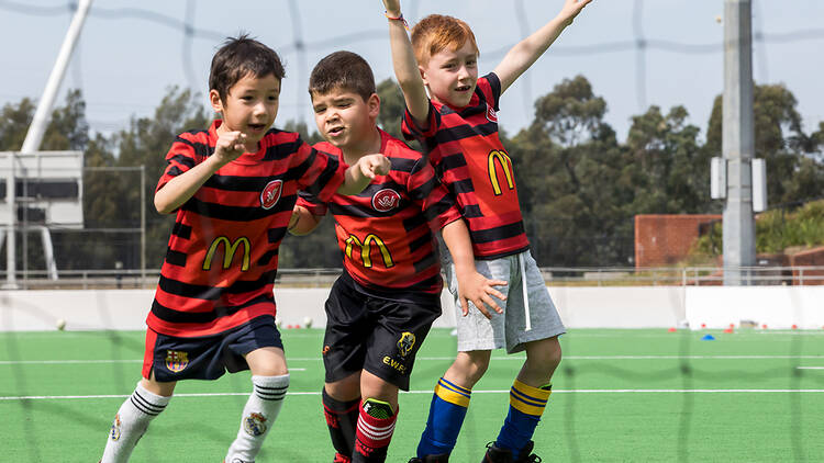 Three boys in red and black soccer uniforms play on a green pitch.