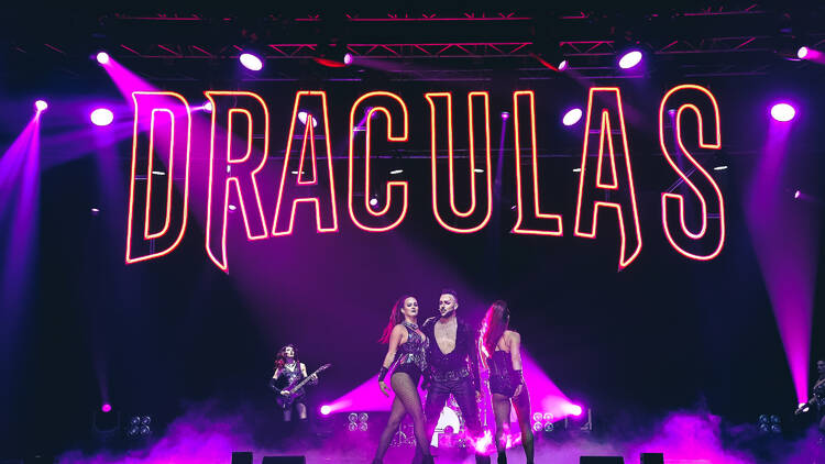 Three people on stage against a yellow neon light reading Dracula's.