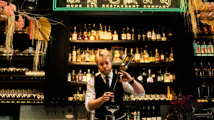 A man in a black vest and tie makes a cocktail at a bar lined with bottles and a green neon sign that says 'Mongkok Restaurant Company'