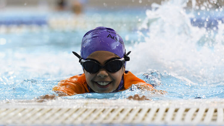 A child in a purple swim cap and goggles kicks in the pool.