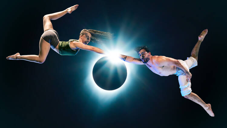 Two circus performers are suspended in darkness, both reaching toward an illuminated eclipsed moon