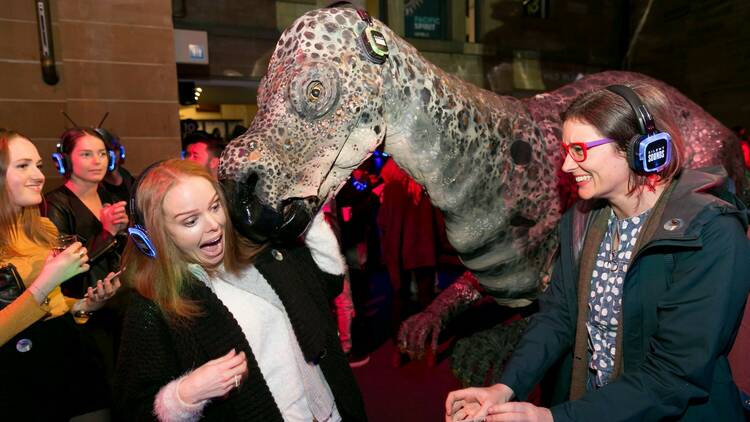 Two women wearing headphones pretend to be scared by a dinosaur statue.