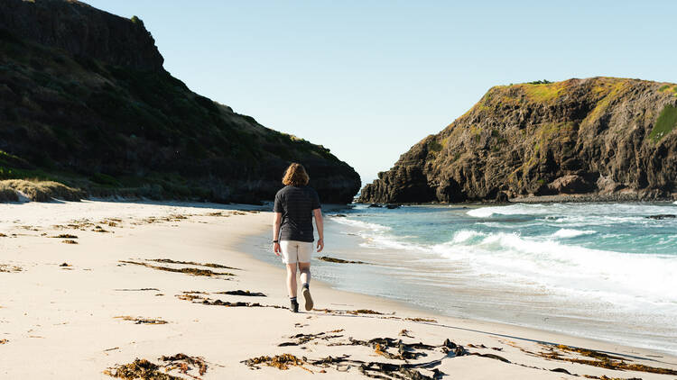 A person walks along a white-sand beach strewn with seaweed.