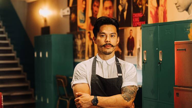 A man with a moustache and wearing a black apron stands with his arms folded in a room filled with posters and lockers.