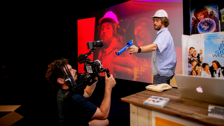 In a play a man wears a hard hat and aims a drill at a man holding  a film camera
