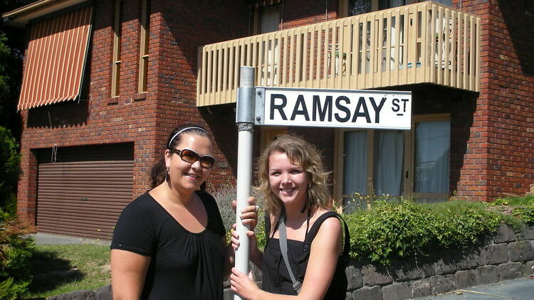 Two women posing in front of a house by a sign that says Ramsay St.