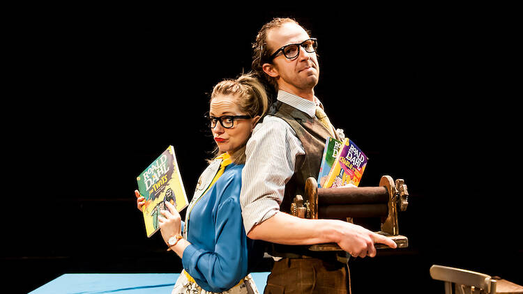 A man and a woman both wearing glasses stand back-to-back holding some books.