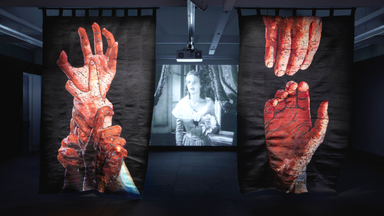 Two tapestries of hands reaching hang from a ceiling, a black and white movie plays on a screen behind them
