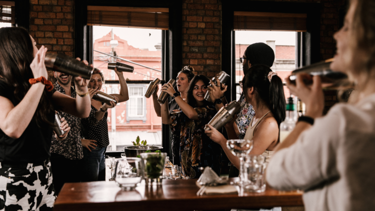 A group of women stand around a table shaking cocktail mixers.