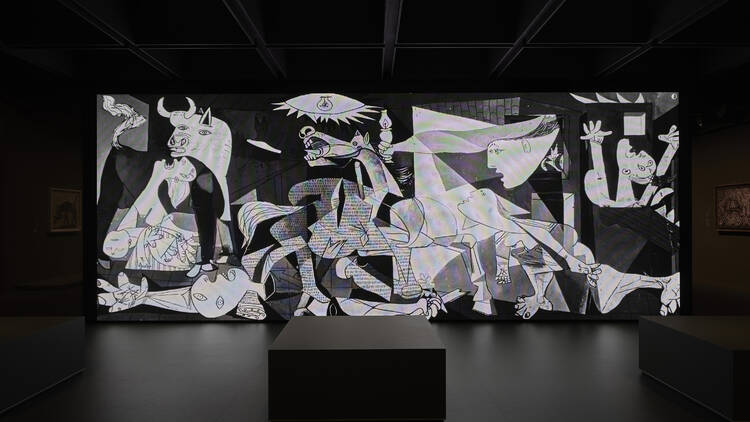 A black and white depiction of Guernica dominates a dark room in the gallery