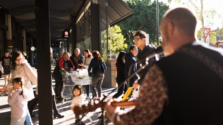 People walk around a busker in late afternoon light at the Marrickville Metro