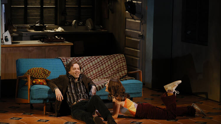 A man and woman sit on the floor of an apartment, leaning against a blue couch