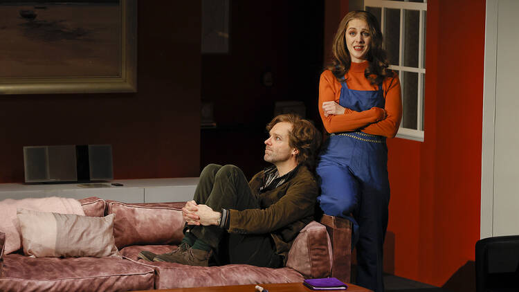 A man sits on a couch, facing away from a woman looking at the audience. She looks unimpressed.