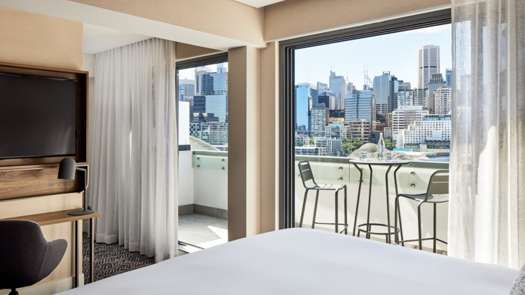 A shot of a white bed corner and open sliding doors opening out to a view of Darling Harbour at the Aiden Darling Harbour 