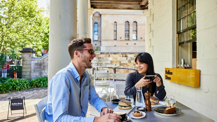 Two customers sit at a table on the verandah of the Mint, enjoying food and drink from the Bullion Store.