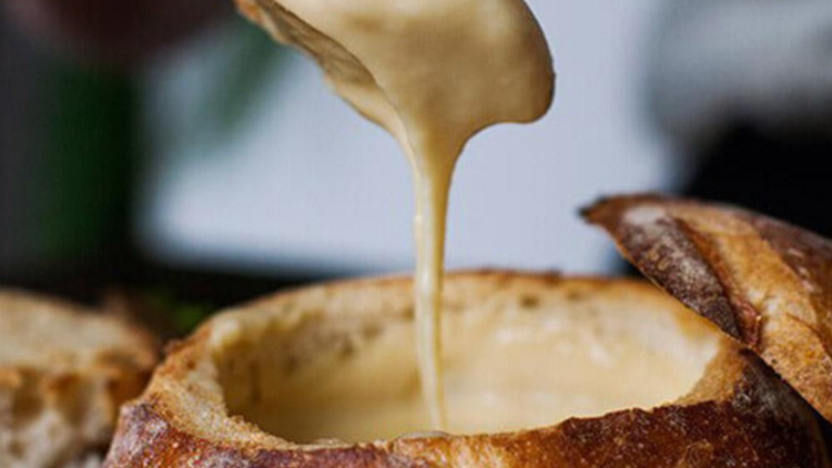 a piece of cheesy bread drips with melted cheese above a crusty bread bowl 