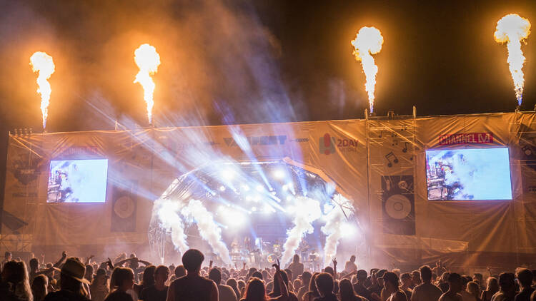 An outdoor music festival stage with big screens, flames and lights, and a huge crowd watching on.