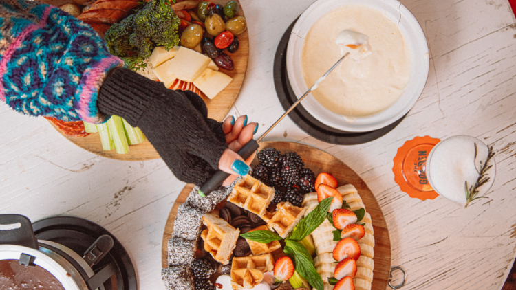 Overhead shot of a table covered in melted cheese fondue and waffles with one hand in a glove dipping into the cheese