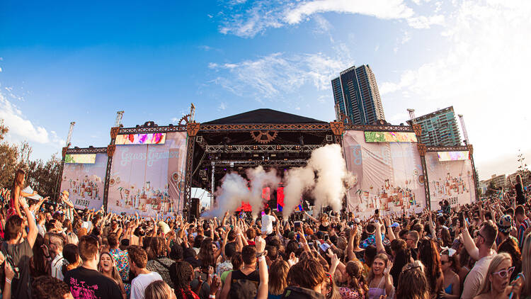 A festival crowd dance with their hands in the air against a stage with plumes of smoke 