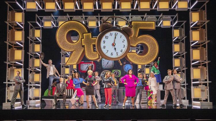 The cast of 9 to 5 the musical pose on stage underneath a huge sign that says '9 to 5' adorned by a giant alarm clock