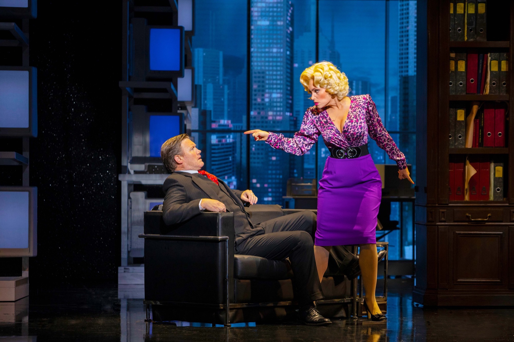 A woman in purple points and stands over a businessman sitting in a chair