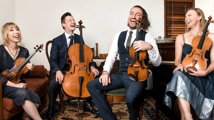 Four musicians sit together laughing while holding their string instruments.