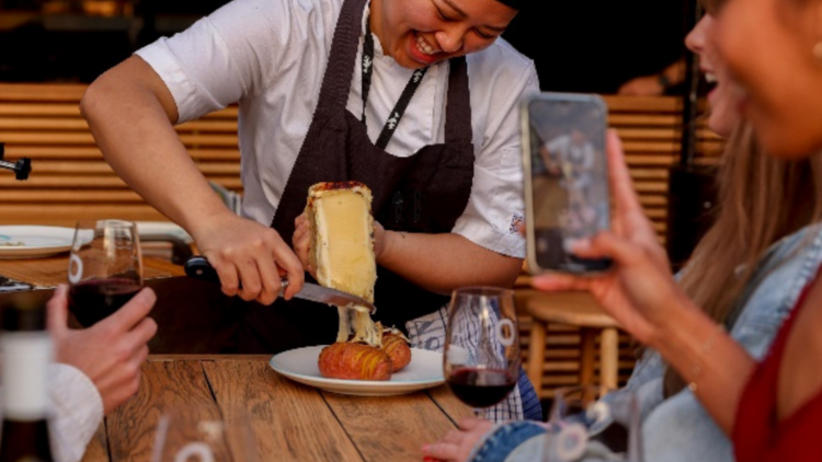 A chef scrapes melting raclette onto a plate at Opera Bar with people taking photos in front