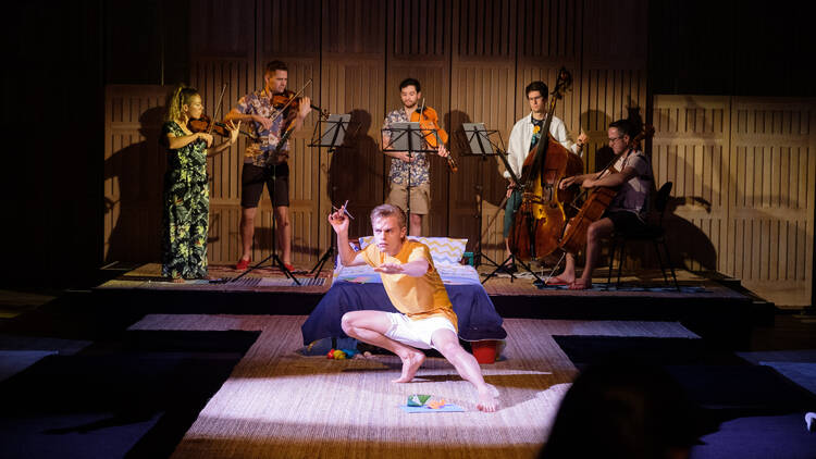 A man in a yellow t-shirt crouches on a stage under the spotlight with a band behind him.