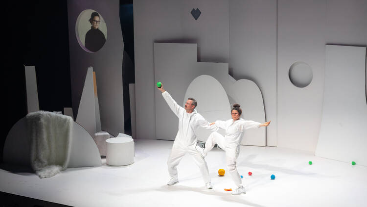 A man and a woman all dressed in white pose with their arms outstretched on an all-white stage dotted with some small coloured balls.