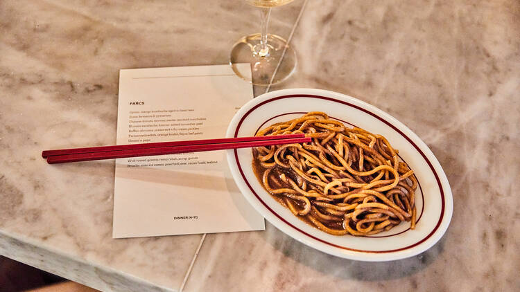 A dish of noodles atop a menu and flanked by a glass of wine.