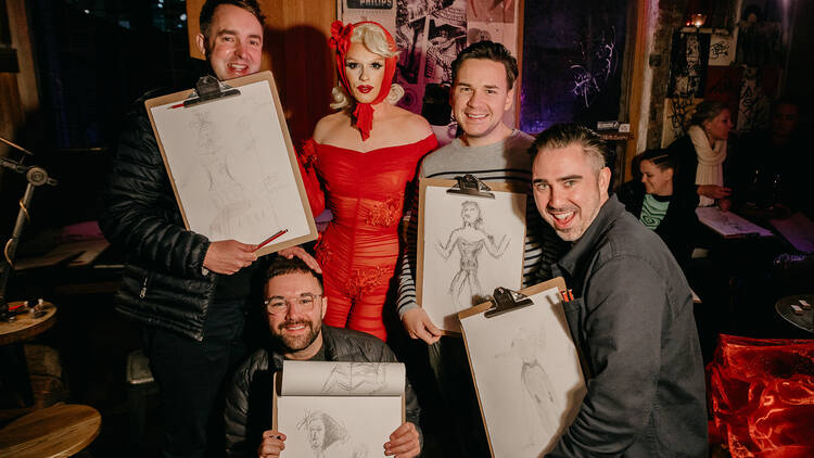 Four men with hand drawn pictures surround drag queen Aubrey Haive, dressed in a red outfit and scarf