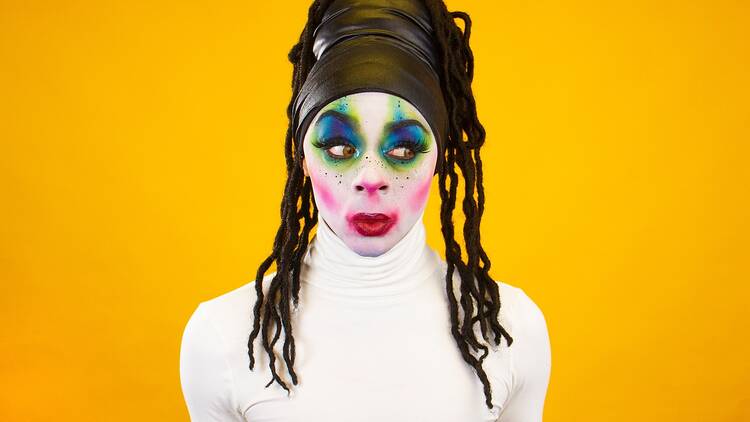 Against a bright yellow background a person in a white top has a face covered in very colourful make-up