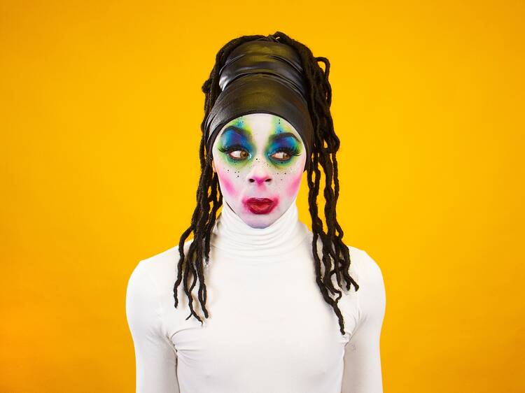 Against a bright yellow background a person in a white top has a face covered in very colourful make-up