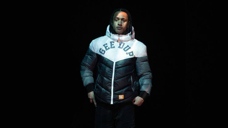 Hip-hop artist Blueboy wears a black and white puffer jacket that says 'geed up' while standing against a black backdrop.