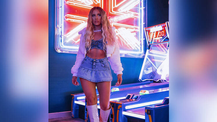 Singer Amarni wears a two-piece denim ensemble with white fluffy jacket in front of a neon-lit Union Jack.
