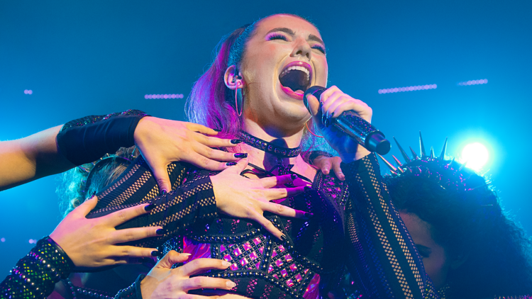 A brunette woman with a ponytail and wearing a pink and black two-piece period costume sings passionately into a microphone while hands grab at her body.