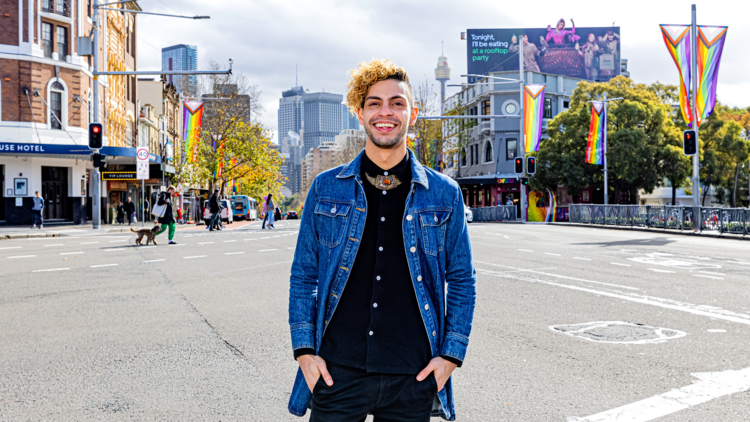 AJ stands at the intersection of Taylor Square with progress pride flags and the busy street behind him he wears a long denim coat over al all black outfit