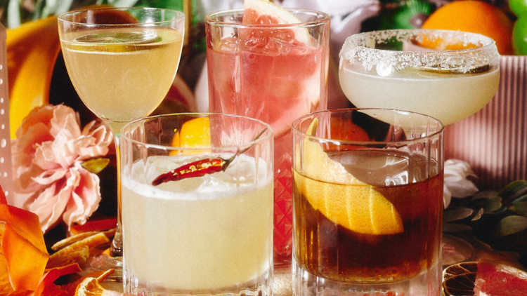 Five colourful tequila cocktails sit against a backdrop of flowers and fruit peel