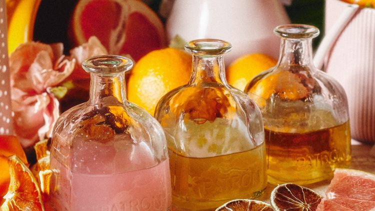 Three glass squat tequila bottles filled with pink and amber liquid sit next to each other against a backdrop of flowers and fruit peel