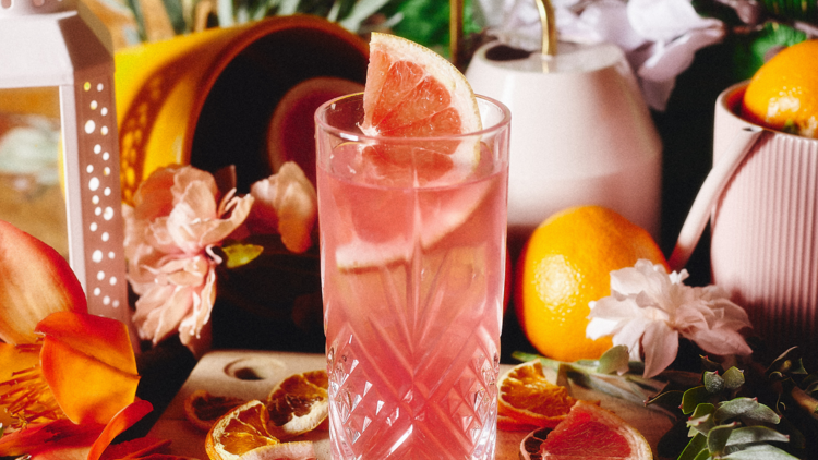 A pink coloured tequila cocktail sits against a backdrop of flowers and fruit peel