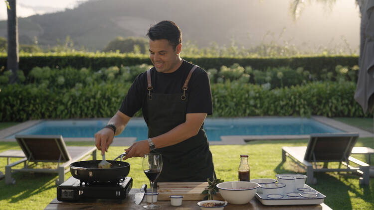 A man in an apron cooks over a camp stove in a garden with a pool.