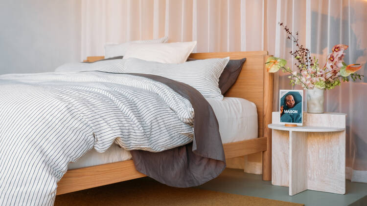 A bed on a wooden base with striped blue and white bedding next to a side table with colourful flowers.
