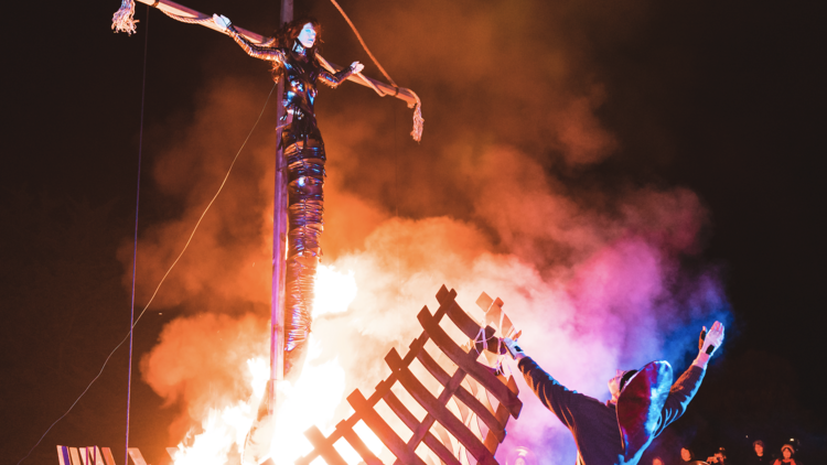 A man stands in front of a burning statue with his arms raised to the sky as people watch from the sidelines.