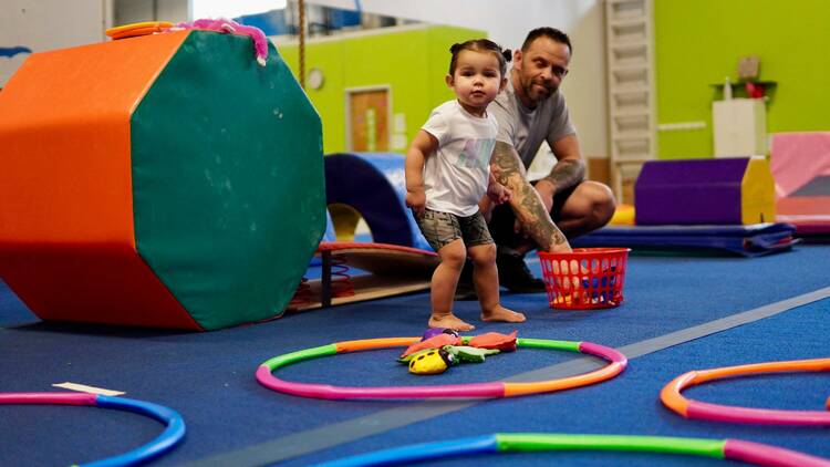 A toddler plays with hula hoops and bean bags with her dad at an indoor play centre.
